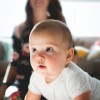 A baby trying to crawl while looking ahead. The baby's mum is out of focus in the background of the image.