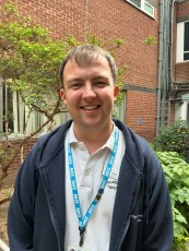 Alex is stood outside and is wearing a uniform of a polo shirt and hoodie.