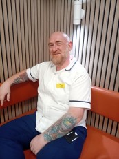 David is sat down and is wearing a uniform of a white clinical tunic.