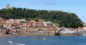 Panoramic view of Scarborough seafront with the harbour, sandy beach, and Scarborough Castle on the green headland overlooking the town.