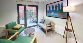 Bright seating area with green and grey chairs, a small coffee table, a floor lamp and large landscape artwork, with glass doors opening onto a garden courtyard.