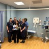 Group of NHS healthcare staff standing in a clinical examination room with a patient holding an information booklet, smiling towards the camera.