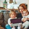 A mum with a ginger bob haircut sat on a grey sofa with a toddler holding an iPad and a baby on her lap as they all watch something on the iPad screen