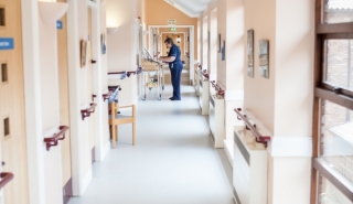 Looking down a long bright hospital corridor with treatment rooms on the left and windows on the right. At the bottom of the corridor is one member of staff in a blue nursing uniform