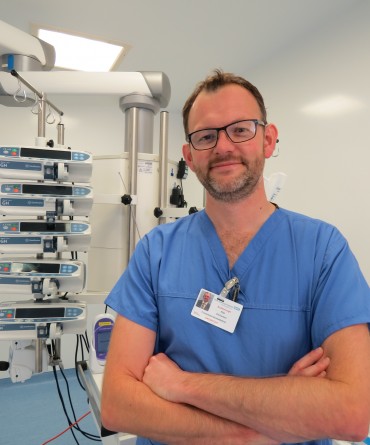Phil in his blue scrubs, standing in front of medical equipment in a hospital room.