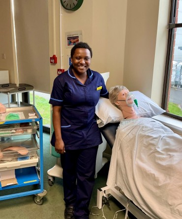 Liz in her dark blue nurse's uniform, standing alongside a medical teaching manikin in a hospital bed.