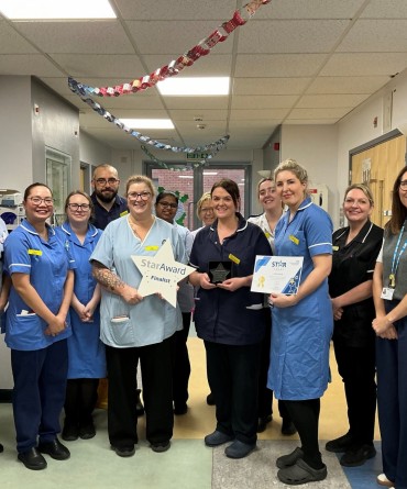 Star Award winners, Ash Ward, receiving their award from Clare Smith, Chief Executive. The team are in nursing uniforms and stood in the corridor of a hospital ward.