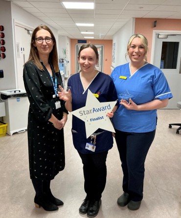 Star Award winner Hannah Davis and Gemma Morris receiving her award from Chief Executive, Clare Smith. Hannah and Gemma are wearing nursing scrubs and they are stood on a hospital ward's corridor.