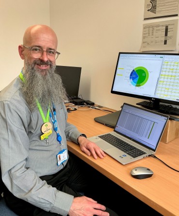 Smiling man with a long beard sits at a desk with a laptop, showing data on the screen. A monitor beside him displays a colourful pie chart. He wears an NHS identify badge.