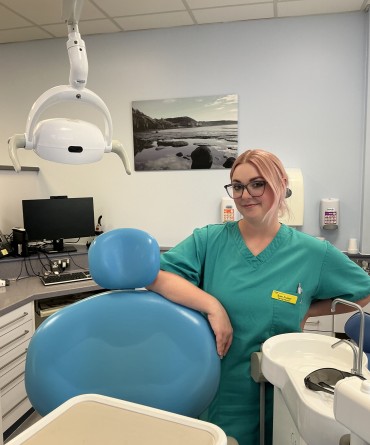 A nurse in turquoise scrubs stood behind a dental chair.