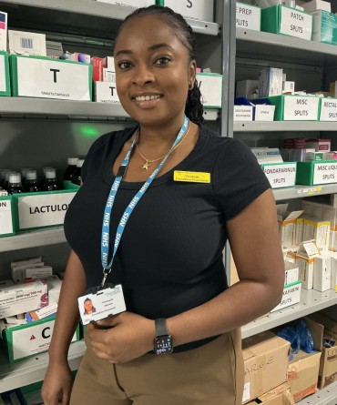A pharmacy staff member stands in a medicines storeroom in front of shelves stocked with medications. The staff member is wearing an NHS lanyard, identification badge and name badge.