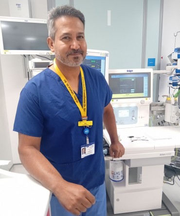 Consultant anaesthetist wearing blue scrubs standing beside anaesthetic and patient monitoring equipment in a hospital clinical room.
