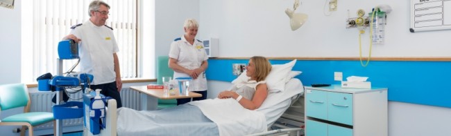 Two members of staff speaking to a patient recovering in a hospital bed.