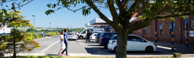 Banner image featuring two patients walking towards a hospital entrance. There are cars and an ambulance parked outside the entrance.