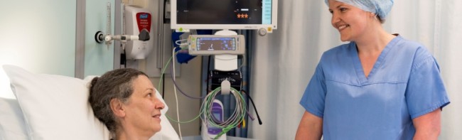 Banner image of a patient in a hospital bed, with a healthcare professional standing alongside them, wearing blue scrubs and a surgical cap. The patient is hooked up to an obs machine which shows their heart rate as 96.