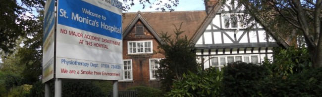 The front of St Monicas hospital from outside with a sign for the hospital on the lawn