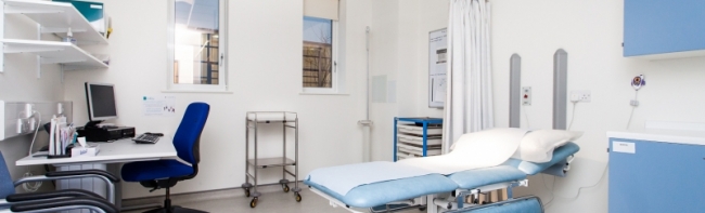 An empty clinical room with a hospital bed, chair and desk