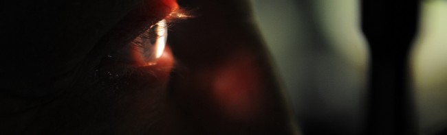 A close up view of a person's side profile. Everything is dark apart from the eye which is lit by an external light source. Used as a page header.