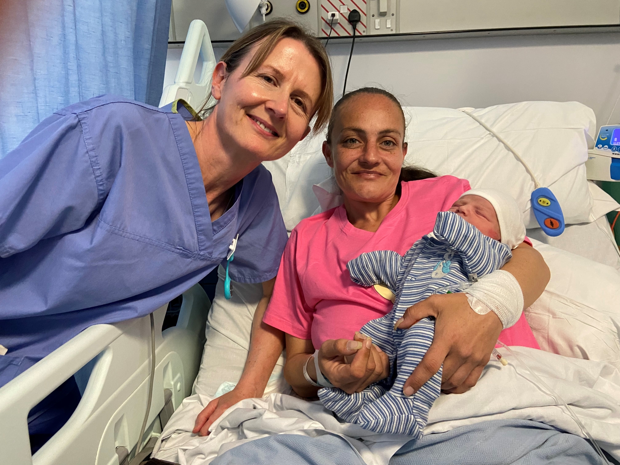 Research Midwife Marie Home posing with new mum Danielle Woodward and her baby son Leif. Danielle is sitting in a hospital bed, holding Leif, while Marie stands alongside them in her work scrubs.